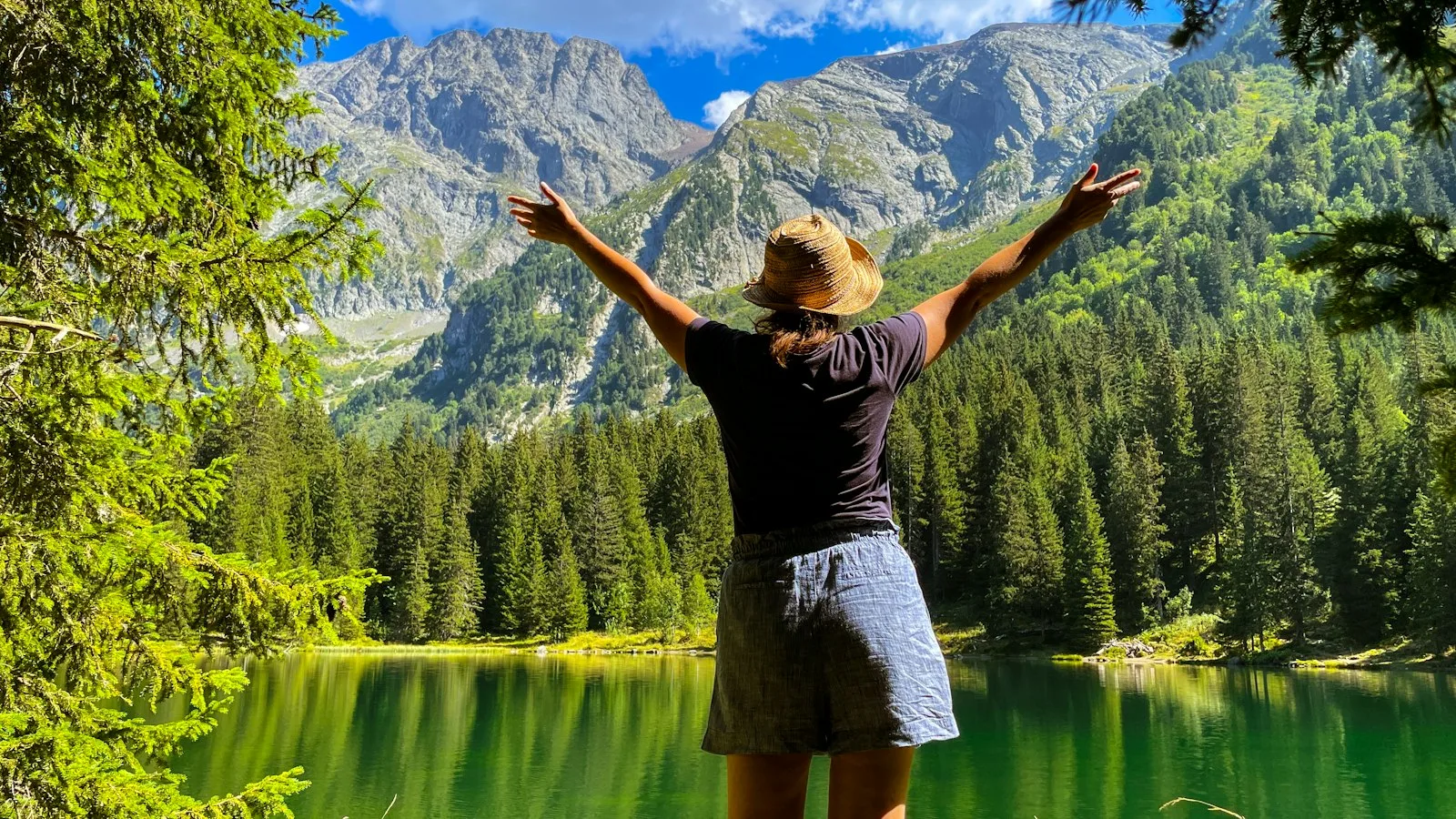 Une voyageuse, dos à la caméra, bras ouverts face à un lac de montagne entouré de forêts dans les Alpes françaises — la promesse de ne rien renoncer.