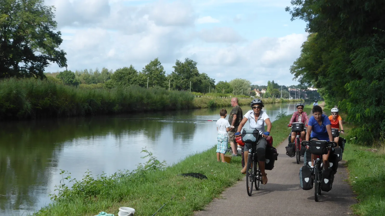 Voyage à vélo en France : un petit groupe de cyclotouristes chargés de sacoches roule sur un chemin de halage le long d'un canal, en été.